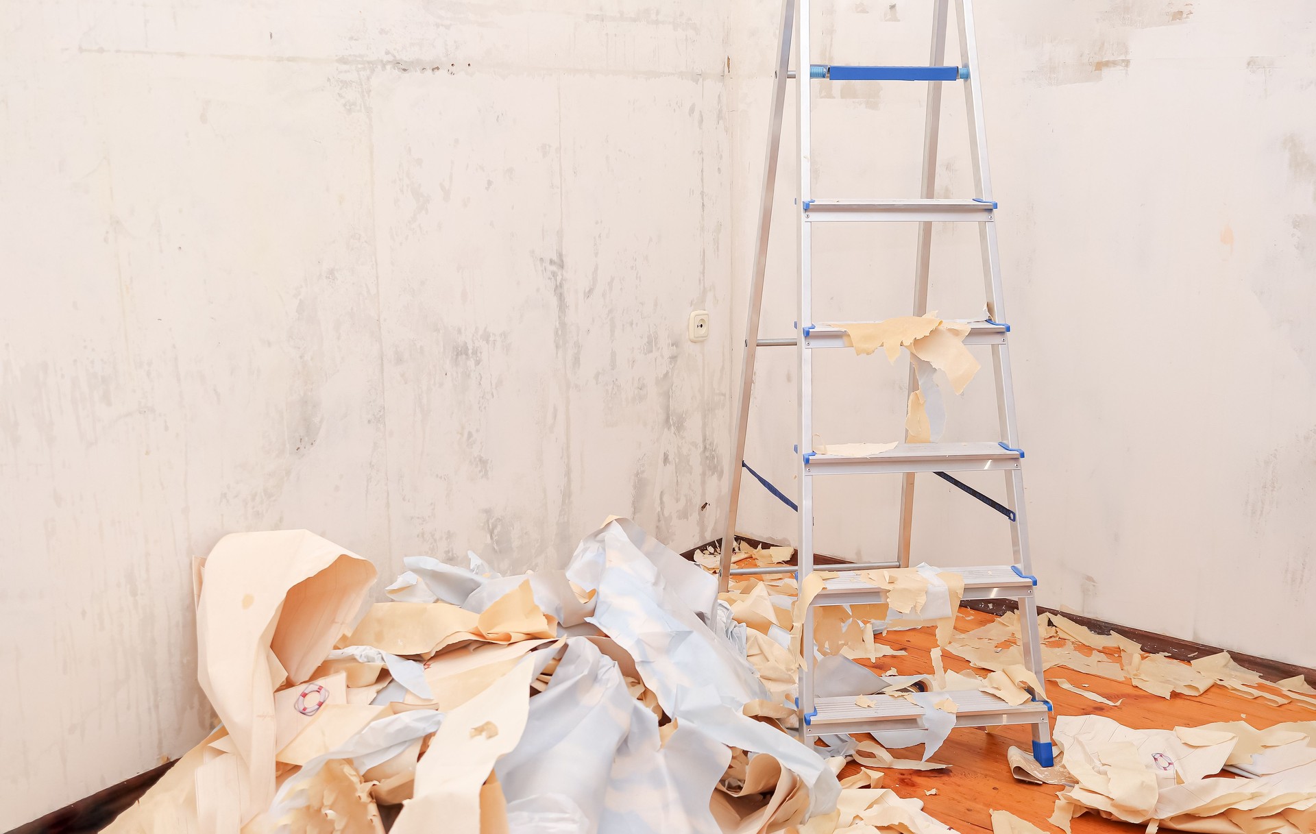 a stepladder stands in the middle of an empty room with old wallpaper removed from the walls. wallpaper is lying on the floor, trash and dust during renovation